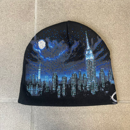 Supreme FW26 Cityscape Art Dealer Black Beanie is a black acrylic beanie with original artwork by Art Dealer, featuring a blue and white night city graphic with a moon and small stars across the sky on both sides. An old English-style Supreme logo sits on the front, and rhinestone appliqués in different sizes decorate the front and back. The print is stretchy, so any cracking effect that appears is just the print stretching with wear.