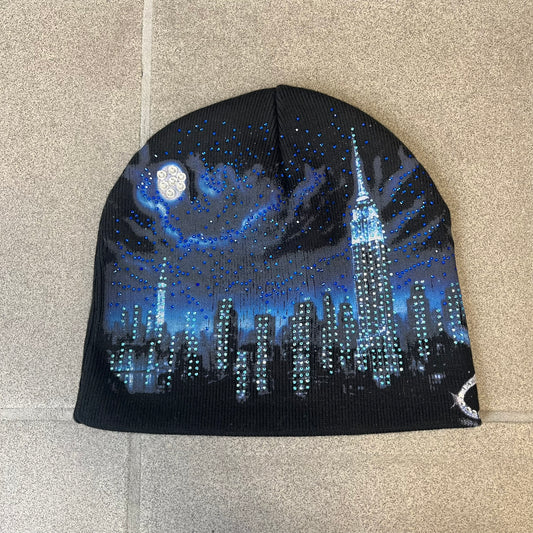 Supreme FW26 Cityscape Art Dealer Black Beanie is a black acrylic beanie with original artwork by Art Dealer, featuring a blue and white night city graphic with a moon and small stars across the sky on both sides. An old English-style Supreme logo sits on the front, and rhinestone appliqués in different sizes decorate the front and back. The print is stretchy, so any cracking effect that appears is just the print stretching with wear.