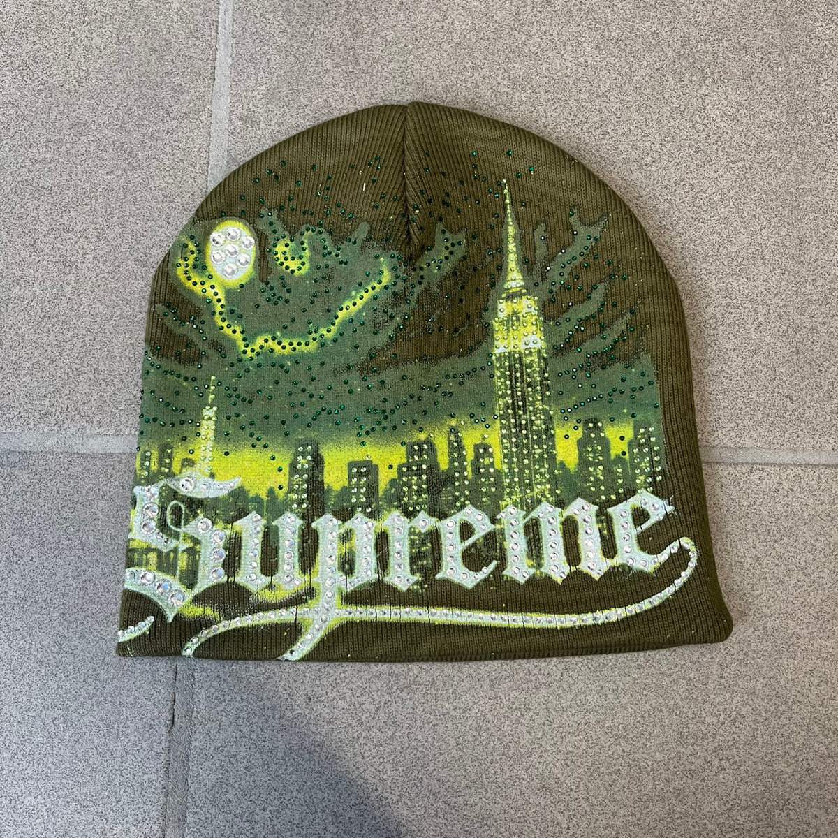 Supreme FW26 Cityscape Art Dealer Green&nbsp; Beanie is a green acrylic beanie with original artwork by Art Dealer, featuring a lime and dark green night city graphic with a moon and small stars across the sky on both sides. An old English-style Supreme logo sits on the front, and rhinestone appliqués in different sizes decorate the front and back. The print is stretchy, so any cracking effect that appears is just the print stretching with wear.