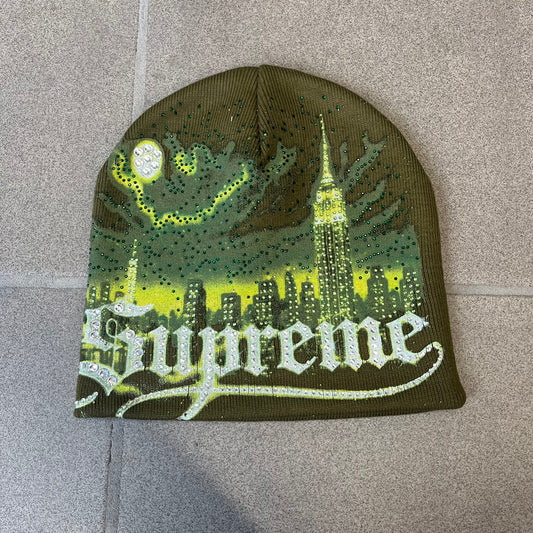 Supreme FW26 Cityscape Art Dealer Green&nbsp; Beanie is a green acrylic beanie with original artwork by Art Dealer, featuring a lime and dark green night city graphic with a moon and small stars across the sky on both sides. An old English-style Supreme logo sits on the front, and rhinestone appliqués in different sizes decorate the front and back. The print is stretchy, so any cracking effect that appears is just the print stretching with wear.