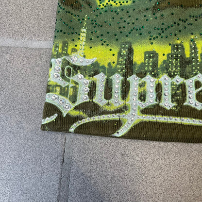 Supreme FW26 Cityscape Art Dealer Green&nbsp; Beanie is a green acrylic beanie with original artwork by Art Dealer, featuring a lime and dark green night city graphic with a moon and small stars across the sky on both sides. An old English-style Supreme logo sits on the front, and rhinestone appliqués in different sizes decorate the front and back. The print is stretchy, so any cracking effect that appears is just the print stretching with wear.