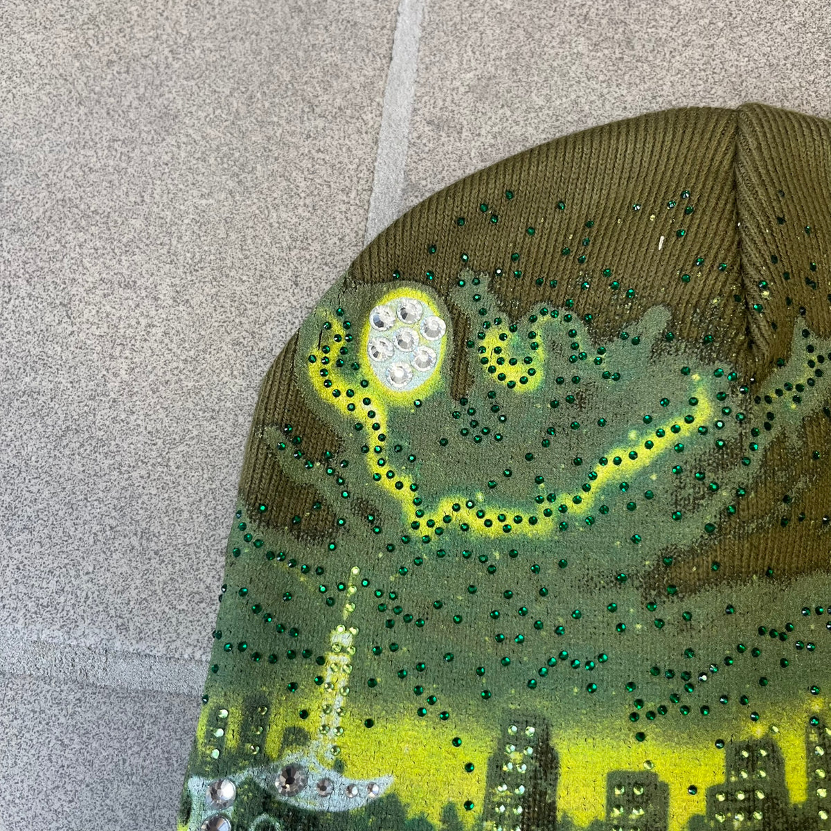 Supreme FW26 Cityscape Art Dealer Green&nbsp; Beanie is a green acrylic beanie with original artwork by Art Dealer, featuring a lime and dark green night city graphic with a moon and small stars across the sky on both sides. An old English-style Supreme logo sits on the front, and rhinestone appliqués in different sizes decorate the front and back. The print is stretchy, so any cracking effect that appears is just the print stretching with wear.