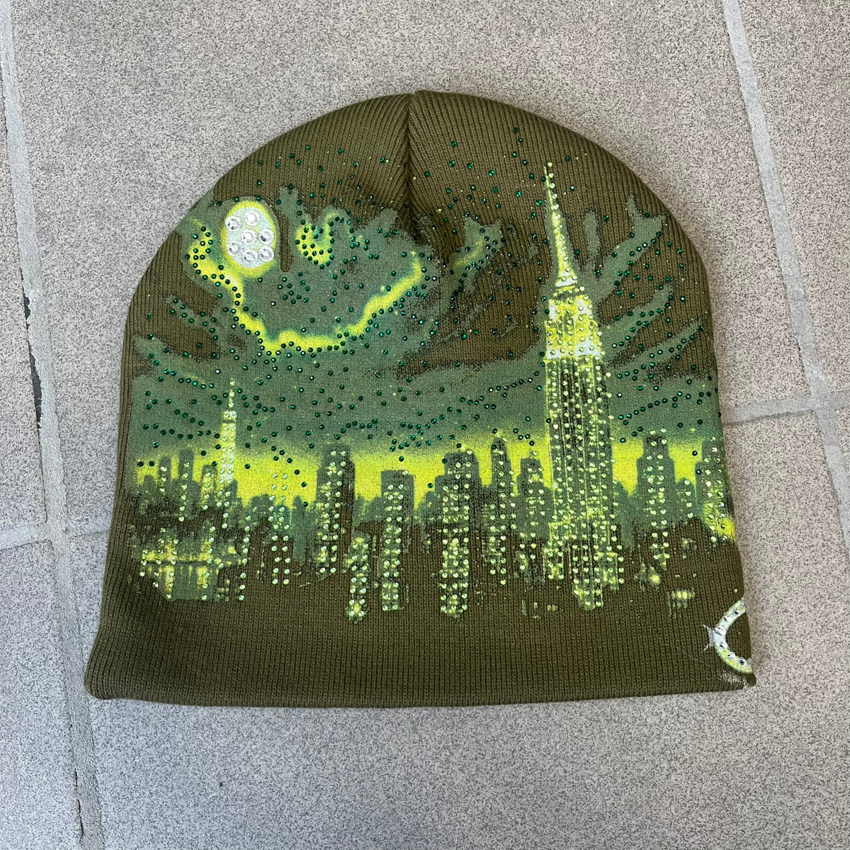 Supreme FW26 Cityscape Art Dealer Green&nbsp; Beanie is a green acrylic beanie with original artwork by Art Dealer, featuring a lime and dark green night city graphic with a moon and small stars across the sky on both sides. An old English-style Supreme logo sits on the front, and rhinestone appliqués in different sizes decorate the front and back. The print is stretchy, so any cracking effect that appears is just the print stretching with wear.