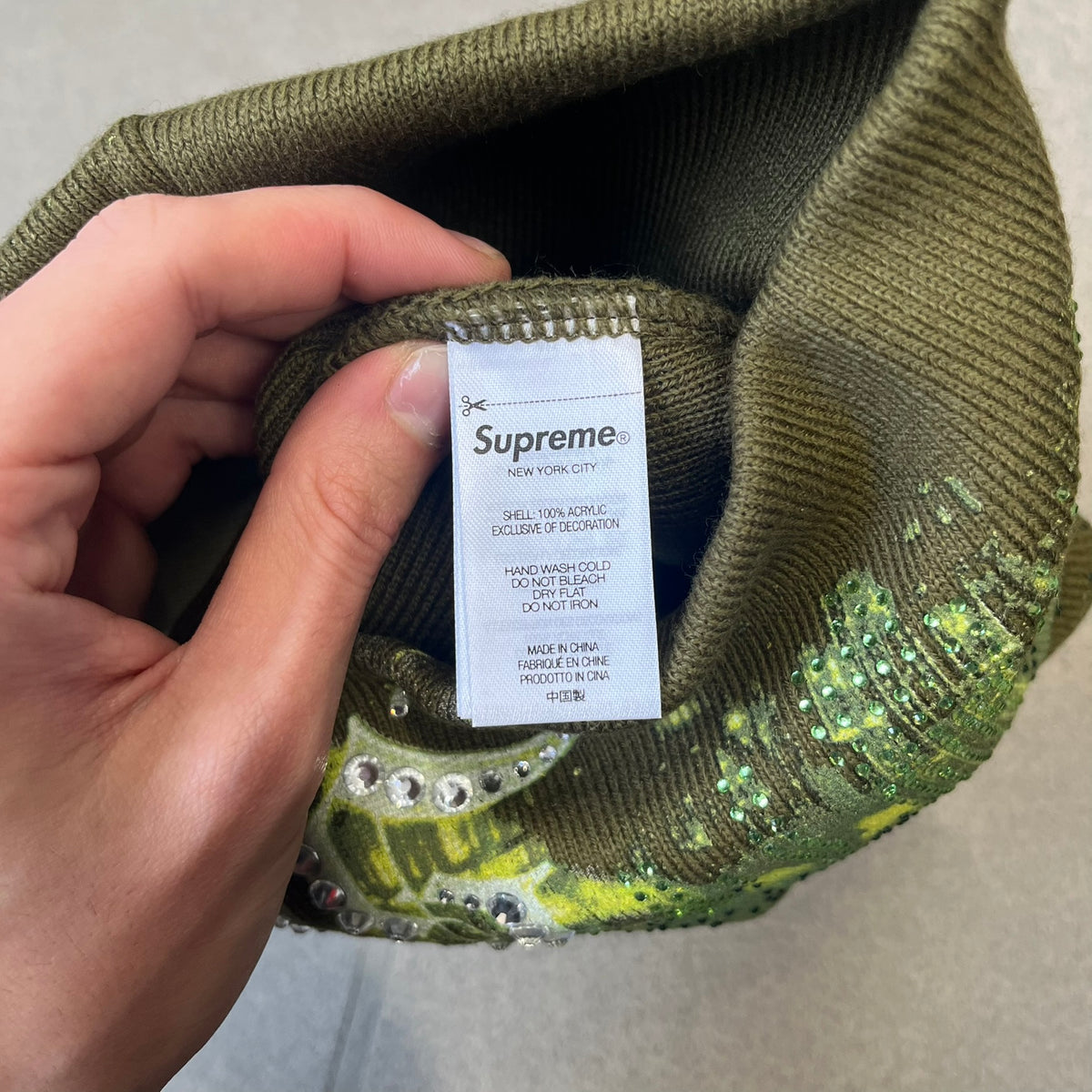 Supreme FW26 Cityscape Art Dealer Green&nbsp; Beanie is a green acrylic beanie with original artwork by Art Dealer, featuring a lime and dark green night city graphic with a moon and small stars across the sky on both sides. An old English-style Supreme logo sits on the front, and rhinestone appliqués in different sizes decorate the front and back. The print is stretchy, so any cracking effect that appears is just the print stretching with wear.