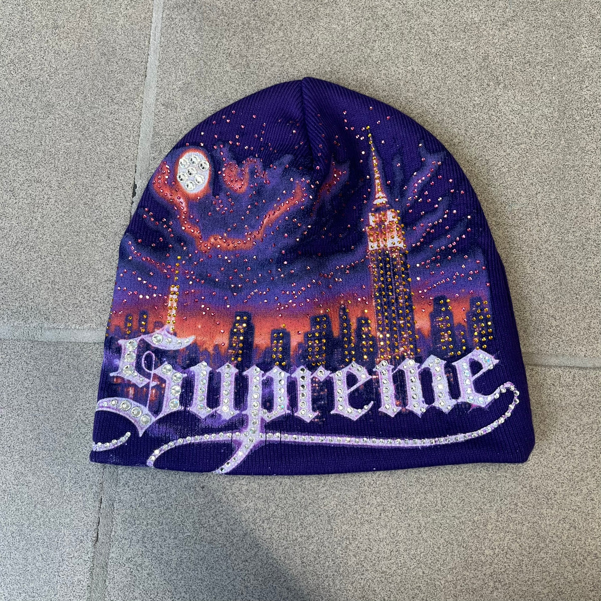 Supreme FW26 Cityscape Art Dealer Purple Beanie is a purple acrylic beanie with original artwork by Art Dealer, featuring a orange and light purple night city graphic with a moon and small stars across the sky on both sides. An old English-style Supreme logo sits on the front, and rhinestone appliqués in different sizes decorate the front and back. The print is stretchy, so any cracking effect that appears is just the print stretching with wear.