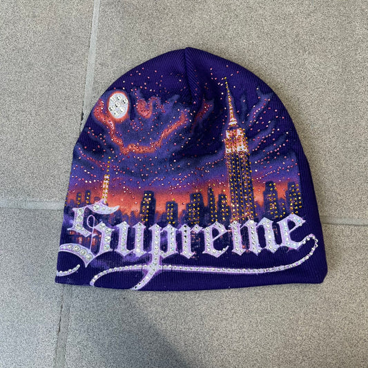 Supreme FW26 Cityscape Art Dealer Purple Beanie is a purple acrylic beanie with original artwork by Art Dealer, featuring a orange and light purple night city graphic with a moon and small stars across the sky on both sides. An old English-style Supreme logo sits on the front, and rhinestone appliqués in different sizes decorate the front and back. The print is stretchy, so any cracking effect that appears is just the print stretching with wear.
