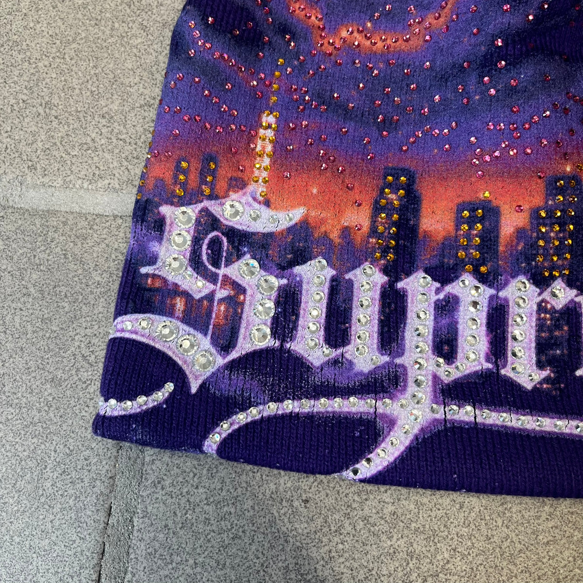 Supreme FW26 Cityscape Art Dealer Purple Beanie is a purple acrylic beanie with original artwork by Art Dealer, featuring a orange and light purple night city graphic with a moon and small stars across the sky on both sides. An old English-style Supreme logo sits on the front, and rhinestone appliqués in different sizes decorate the front and back. The print is stretchy, so any cracking effect that appears is just the print stretching with wear.