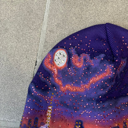 Supreme FW26 Cityscape Art Dealer Purple Beanie is a purple acrylic beanie with original artwork by Art Dealer, featuring a orange and light purple night city graphic with a moon and small stars across the sky on both sides. An old English-style Supreme logo sits on the front, and rhinestone appliqués in different sizes decorate the front and back. The print is stretchy, so any cracking effect that appears is just the print stretching with wear.