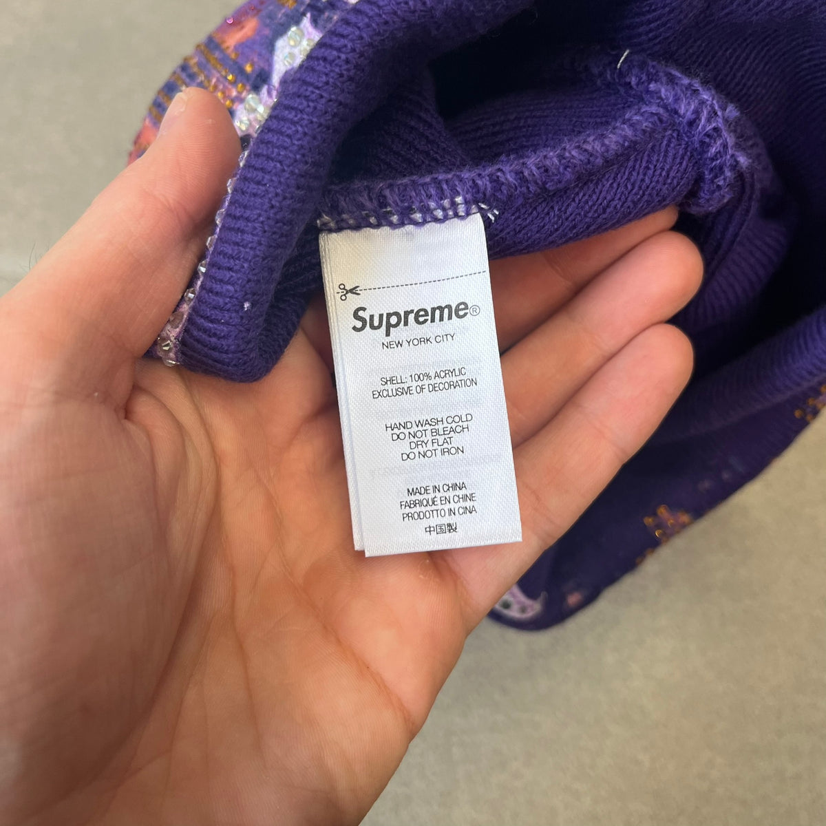 Supreme FW26 Cityscape Art Dealer Purple Beanie is a purple acrylic beanie with original artwork by Art Dealer, featuring a orange and light purple night city graphic with a moon and small stars across the sky on both sides. An old English-style Supreme logo sits on the front, and rhinestone appliqués in different sizes decorate the front and back. The print is stretchy, so any cracking effect that appears is just the print stretching with wear.