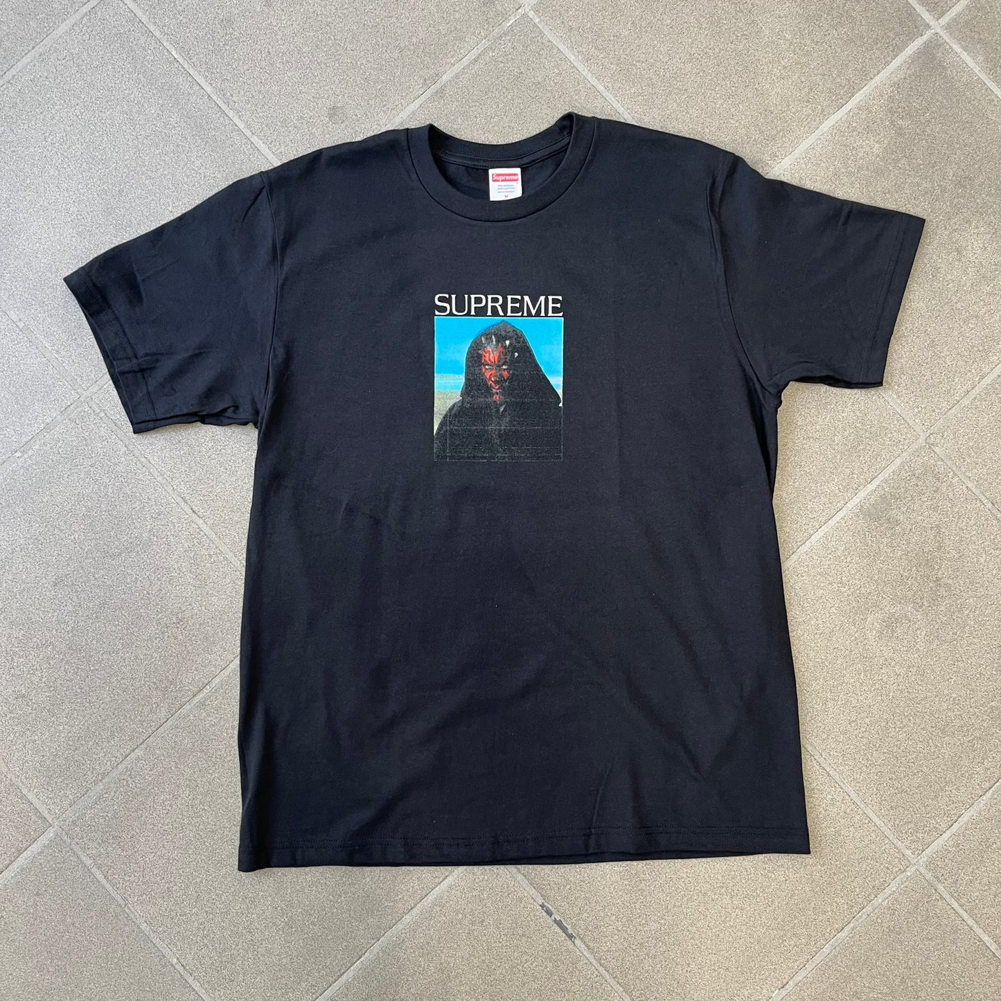 Supreme FW26 Darth Maul Sith Black Tee features a black 100% cotton T-shirt with a photo graphic of Darth Maul on a blue background. A white Supreme logo is printed above the graphic. Features a licensed Star Wars neck tag and is made in Honduras.

