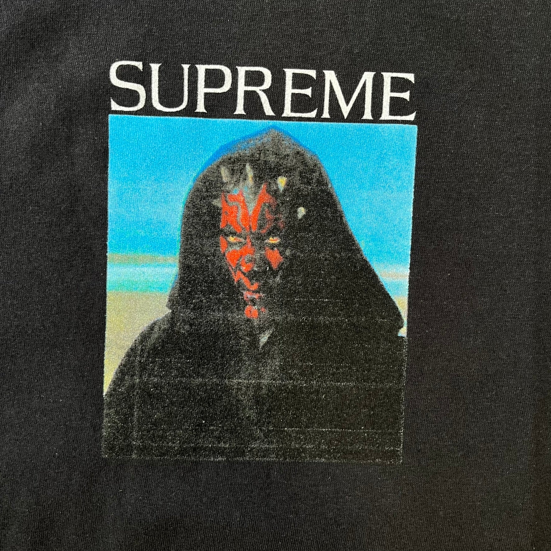 Supreme FW26 Darth Maul Sith Black Tee features a black 100% cotton T-shirt with a photo graphic of Darth Maul on a blue background. A white Supreme logo is printed above the graphic. Features a licensed Star Wars neck tag and is made in Honduras.
