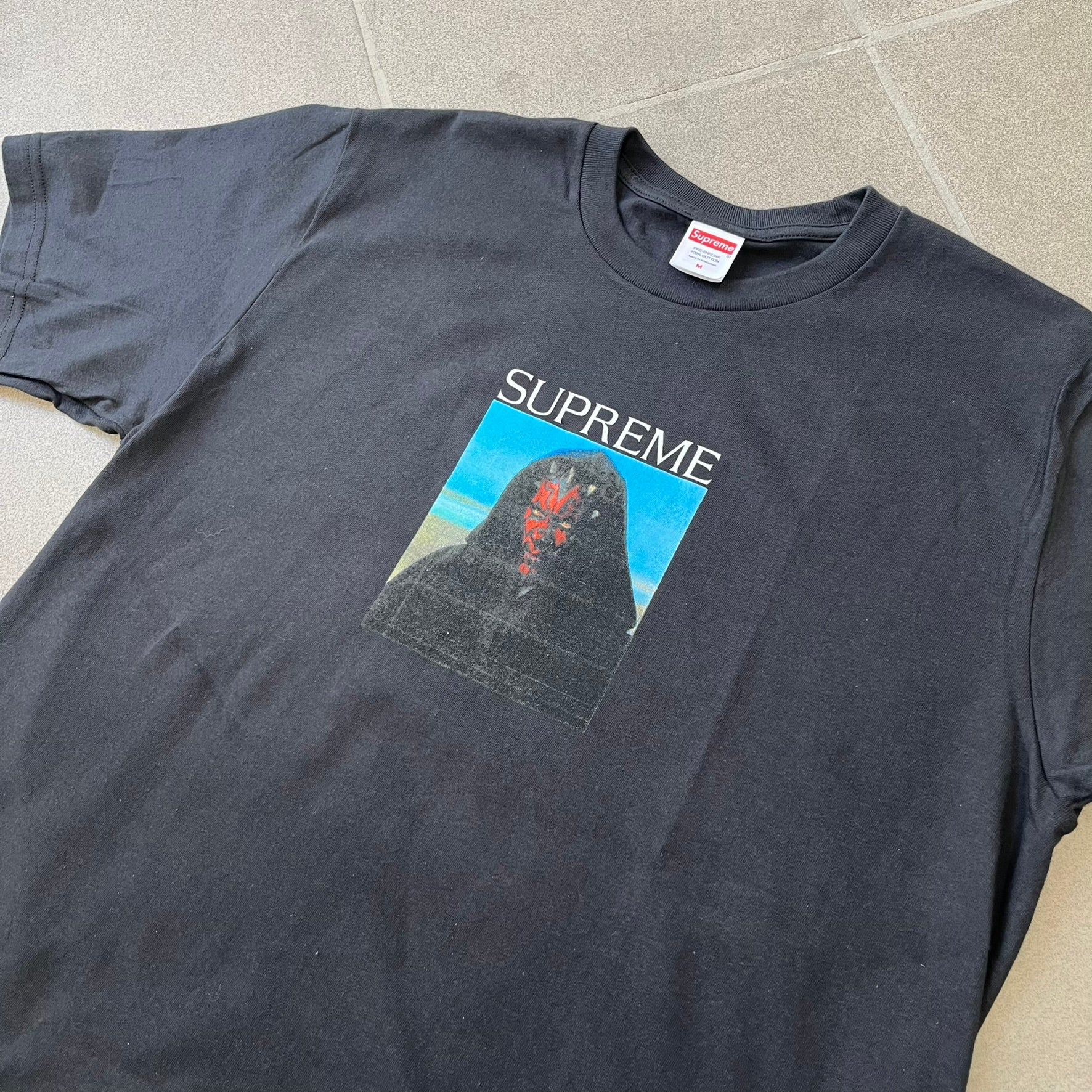 Supreme FW26 Darth Maul Sith Black Tee features a black 100% cotton T-shirt with a photo graphic of Darth Maul on a blue background. A white Supreme logo is printed above the graphic. Features a licensed Star Wars neck tag and is made in Honduras.
