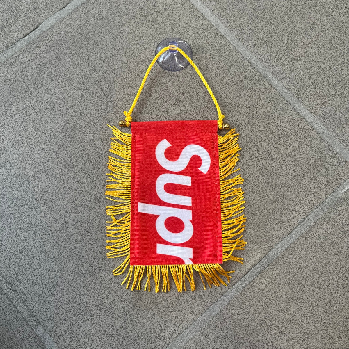 Supreme FW26 Fringed Banner is a small red banner trimmed with yellow fringe along the sides and bottom, with a yellow string at the top for hanging via suction mount. The design features a folded red box logo split across both sides of the banner, and it was distributed as a first-week free gift for FW26.