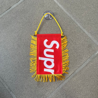 Supreme FW26 Fringed Banner is a small red banner trimmed with yellow fringe along the sides and bottom, with a yellow string at the top for hanging via suction mount. The design features a folded red box logo split across both sides of the banner, and it was distributed as a first-week free gift for FW26.