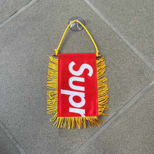 Supreme FW26 Fringed Banner is a small red banner trimmed with yellow fringe along the sides and bottom, with a yellow string at the top for hanging via suction mount. The design features a folded red box logo split across both sides of the banner, and it was distributed as a first-week free gift for FW26.