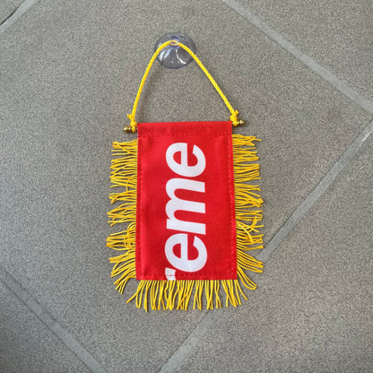 Supreme FW26 Fringed Banner is a small red banner trimmed with yellow fringe along the sides and bottom, with a yellow string at the top for hanging via suction mount. The design features a folded red box logo split across both sides of the banner, and it was distributed as a first-week free gift for FW26.