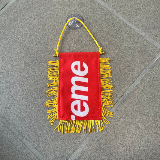 Supreme FW26 Fringed Banner is a small red banner trimmed with yellow fringe along the sides and bottom, with a yellow string at the top for hanging via suction mount. The design features a folded red box logo split across both sides of the banner, and it was distributed as a first-week free gift for FW26.