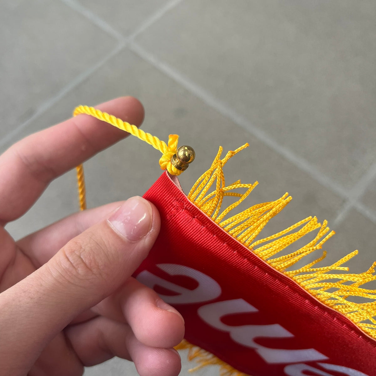 Supreme FW26 Fringed Banner is a small red banner trimmed with yellow fringe along the sides and bottom, with a yellow string at the top for hanging via suction mount. The design features a folded red box logo split across both sides of the banner, and it was distributed as a first-week free gift for FW26.