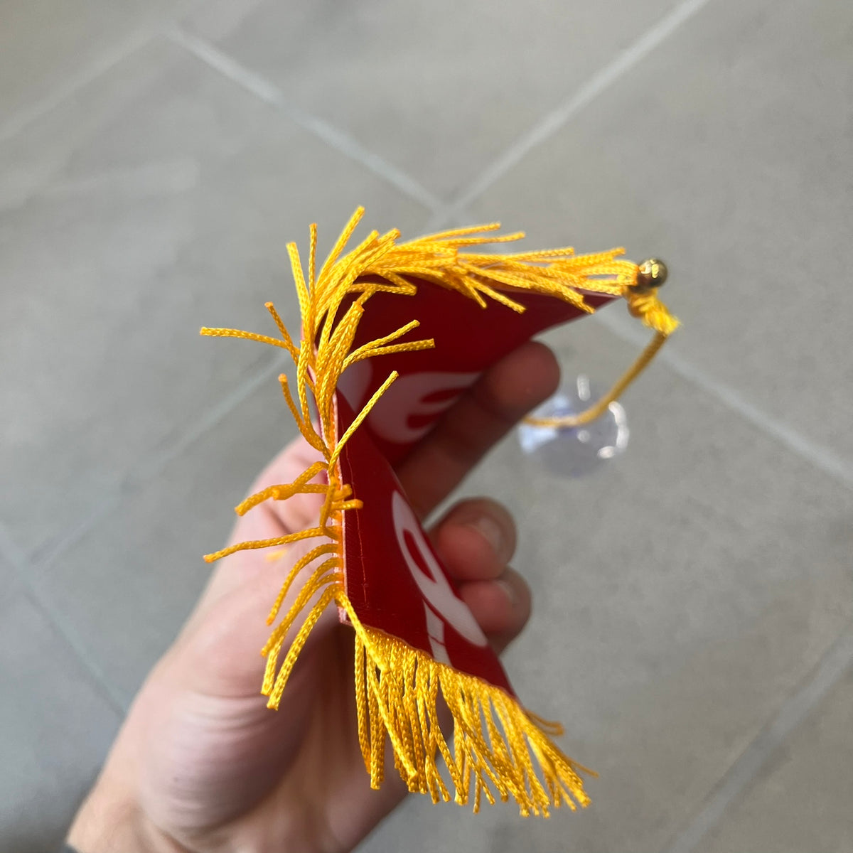 Supreme FW26 Fringed Banner is a small red banner trimmed with yellow fringe along the sides and bottom, with a yellow string at the top for hanging via suction mount. The design features a folded red box logo split across both sides of the banner, and it was distributed as a first-week free gift for FW26.