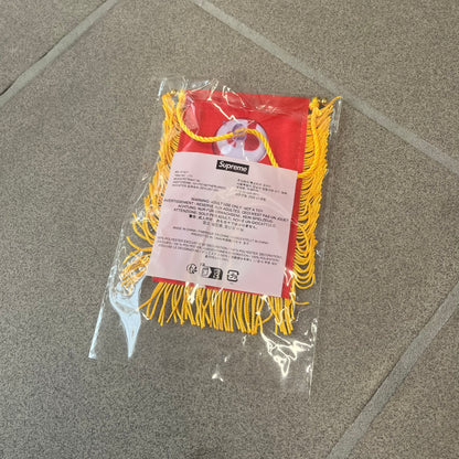 Supreme FW26 Fringed Banner is a small red banner trimmed with yellow fringe along the sides and bottom, with a yellow string at the top for hanging via suction mount. The design features a folded red box logo split across both sides of the banner, and it was distributed as a first-week free gift for FW26.