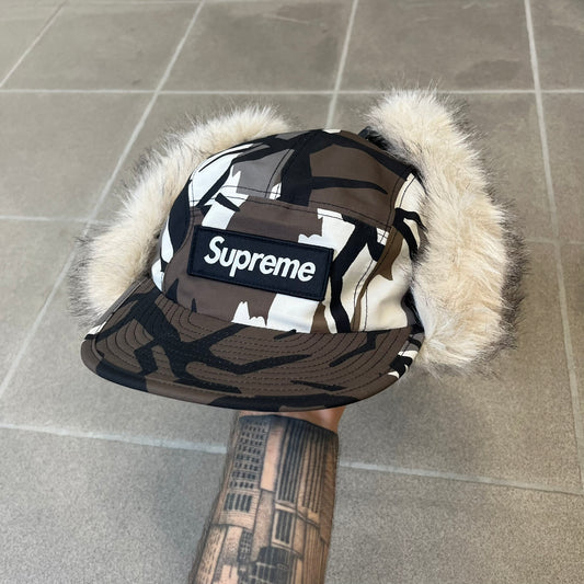 Supreme FW26 GORE-TEX Earflap Predator Camo Camp Cap is a waterproof, breathable camp cap made from GORE-TEX 2-layer poly ripstop in a white, black, and brown camo pattern. It features a black Supreme box logo on the front, an embroidered graphic on the side, and a strap closure. The cap is lined with polar fleece, and the earflaps are lined with faux fur.