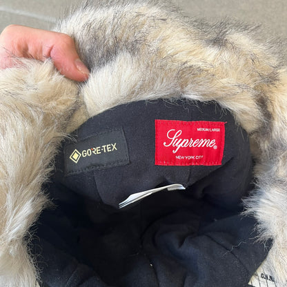 Supreme FW26 GORE-TEX Earflap Predator Camo Camp Cap is a waterproof, breathable camp cap made from GORE-TEX 2-layer poly ripstop in a white, black, and brown camo pattern. It features a black Supreme box logo on the front, an embroidered graphic on the side, and a strap closure. The cap is lined with polar fleece, and the earflaps are lined with faux fur.