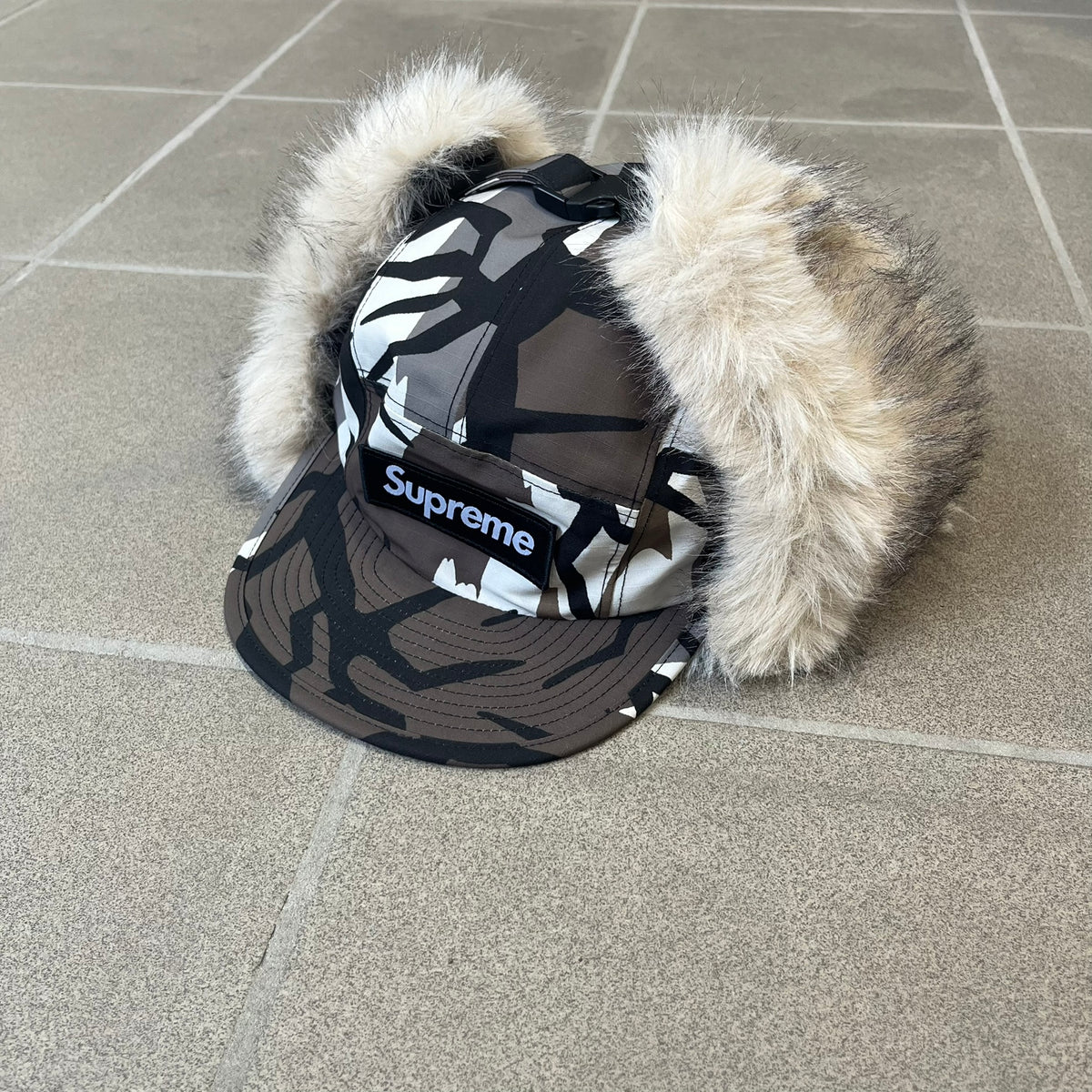 Supreme FW26 GORE-TEX Earflap Predator Camo Camp Cap is a waterproof, breathable camp cap made from GORE-TEX 2-layer poly ripstop in a white, black, and brown camo pattern. It features a black Supreme box logo on the front, an embroidered graphic on the side, and a strap closure. The cap is lined with polar fleece, and the earflaps are lined with faux fur.