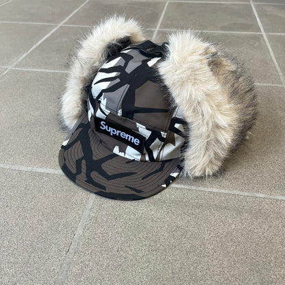 Supreme FW26 GORE-TEX Earflap Predator Camo Camp Cap is a waterproof, breathable camp cap made from GORE-TEX 2-layer poly ripstop in a white, black, and brown camo pattern. It features a black Supreme box logo on the front, an embroidered graphic on the side, and a strap closure. The cap is lined with polar fleece, and the earflaps are lined with faux fur.