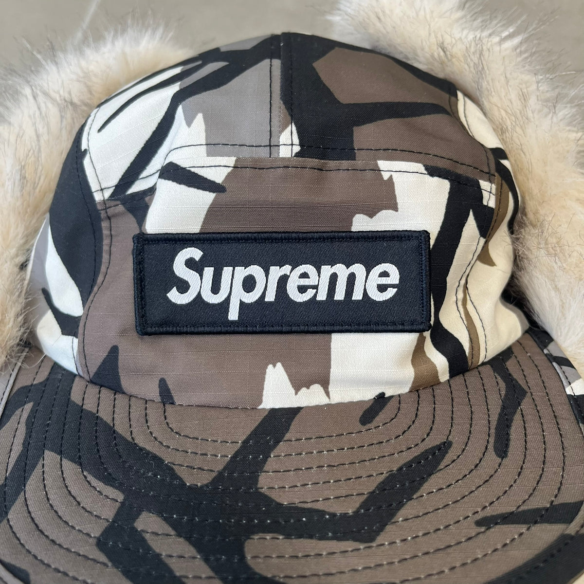 Supreme FW26 GORE-TEX Earflap Predator Camo Camp Cap is a waterproof, breathable camp cap made from GORE-TEX 2-layer poly ripstop in a white, black, and brown camo pattern. It features a black Supreme box logo on the front, an embroidered graphic on the side, and a strap closure. The cap is lined with polar fleece, and the earflaps are lined with faux fur.