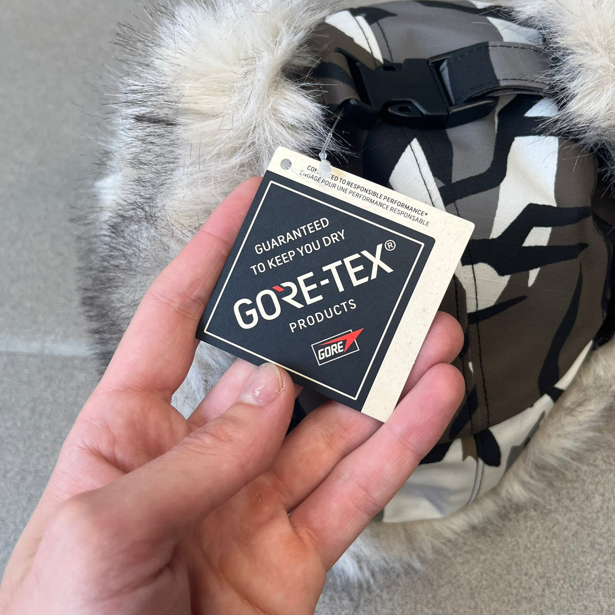 Supreme FW26 GORE-TEX Earflap Predator Camo Camp Cap is a waterproof, breathable camp cap made from GORE-TEX 2-layer poly ripstop in a white, black, and brown camo pattern. It features a black Supreme box logo on the front, an embroidered graphic on the side, and a strap closure. The cap is lined with polar fleece, and the earflaps are lined with faux fur.