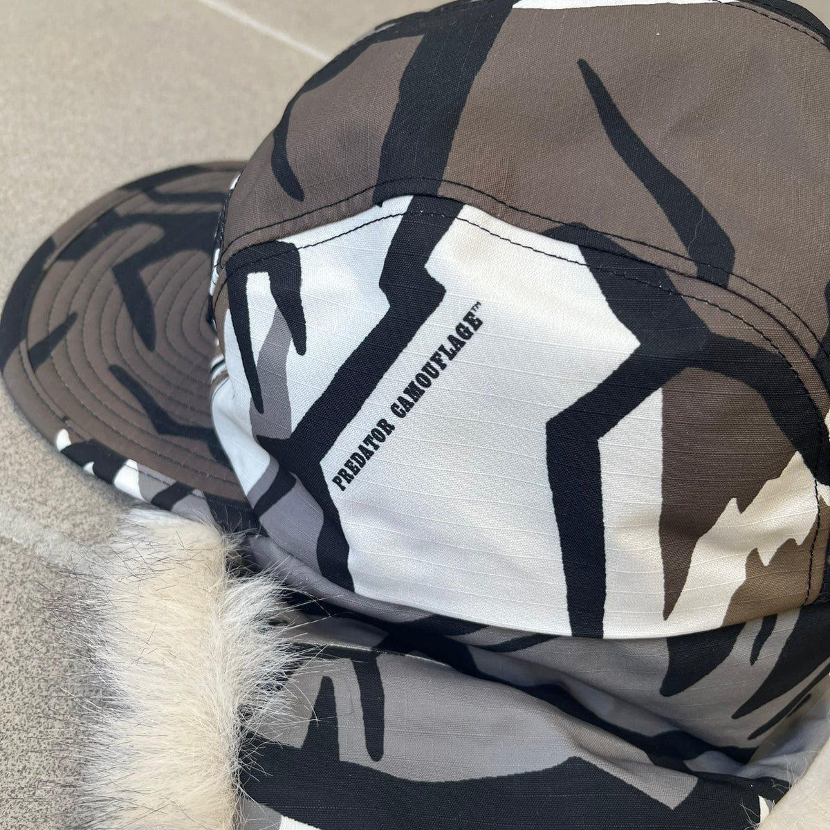 Supreme FW26 GORE-TEX Earflap Predator Camo Camp Cap is a waterproof, breathable camp cap made from GORE-TEX 2-layer poly ripstop in a white, black, and brown camo pattern. It features a black Supreme box logo on the front, an embroidered graphic on the side, and a strap closure. The cap is lined with polar fleece, and the earflaps are lined with faux fur.