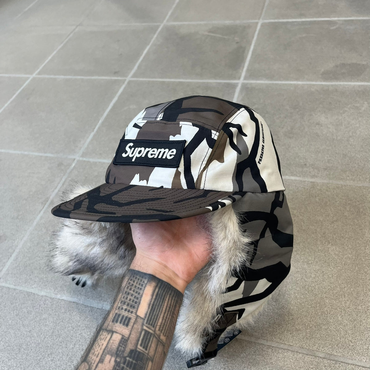 Supreme FW26 GORE-TEX Earflap Predator Camo Camp Cap is a waterproof, breathable camp cap made from GORE-TEX 2-layer poly ripstop in a white, black, and brown camo pattern. It features a black Supreme box logo on the front, an embroidered graphic on the side, and a strap closure. The cap is lined with polar fleece, and the earflaps are lined with faux fur.