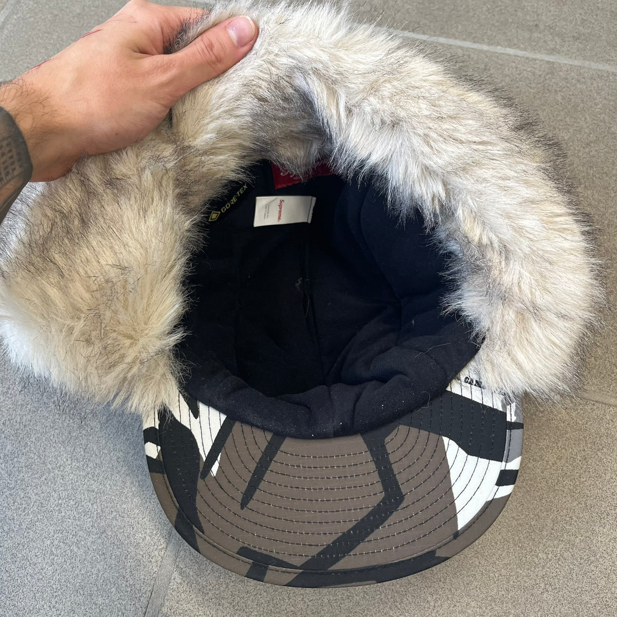 Supreme FW26 GORE-TEX Earflap Predator Camo Camp Cap is a waterproof, breathable camp cap made from GORE-TEX 2-layer poly ripstop in a white, black, and brown camo pattern. It features a black Supreme box logo on the front, an embroidered graphic on the side, and a strap closure. The cap is lined with polar fleece, and the earflaps are lined with faux fur.