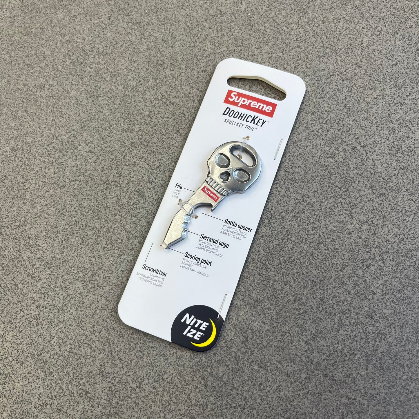 Supreme FW26 Nite Ize Skull Key Tool features a silver-tone multi-purpose key tool with a skull-shaped top and a red Supreme logo underneath. The tool includes a file, bottle opener, serrated edge, scoring point, and screwdriver.
