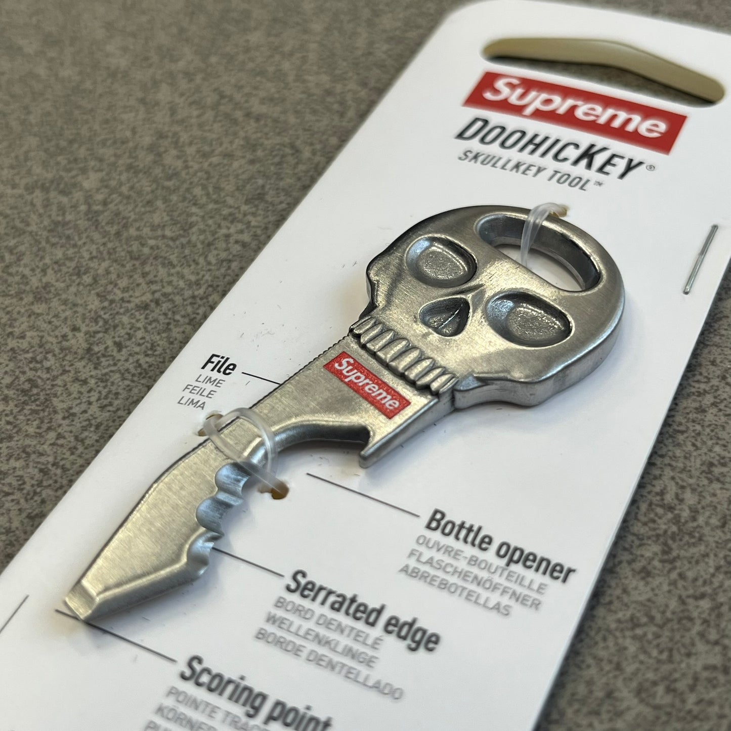 Supreme FW26 Nite Ize Skull Key Tool features a silver-tone multi-purpose key tool with a skull-shaped top and a red Supreme logo underneath. The tool includes a file, bottle opener, serrated edge, scoring point, and screwdriver.
