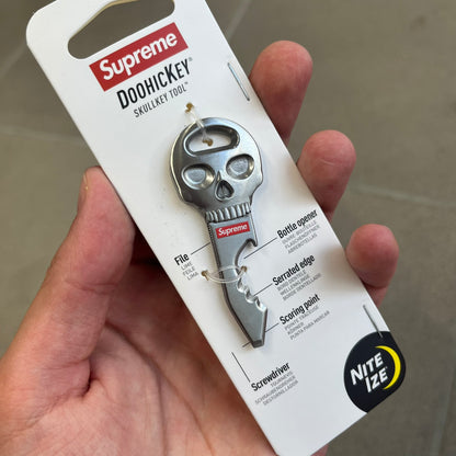 Supreme FW26 Nite Ize Skull Key Tool features a silver-tone multi-purpose key tool with a skull-shaped top and a red Supreme logo underneath. The tool includes a file, bottle opener, serrated edge, scoring point, and screwdriver.
