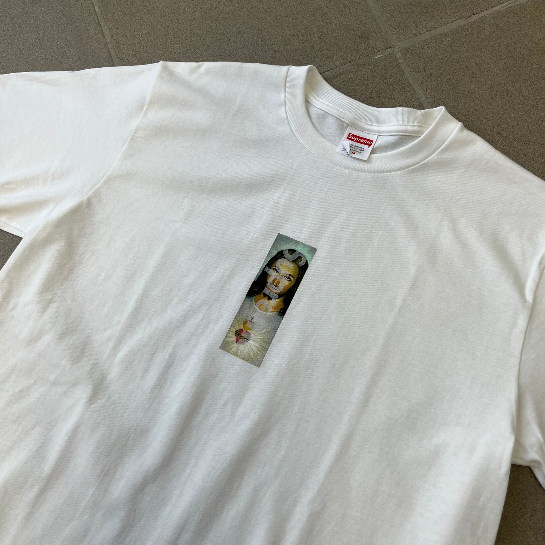 Supreme FW26 Prayer Card Jesus Box Logo White Tee features a white 100% cotton T-shirt with a vertical Supreme box logo graphic on the front. The Supreme text is printed lightly over a graphic depicting the Sacred Heart of Jesus. Made in Honduras.
