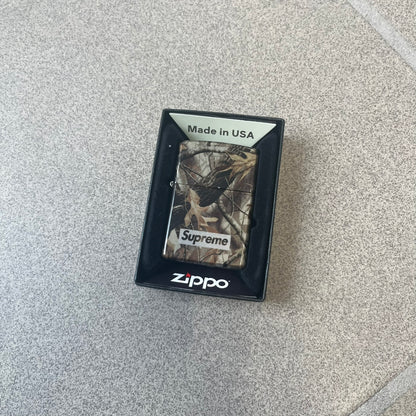 Supreme FW26 RealTree Camo Logo Zippo Lighter is an original Zippo featuring a full RealTree camo graphic with the Supreme box logo on the front. It comes with the original Zippo box and has never been filled.