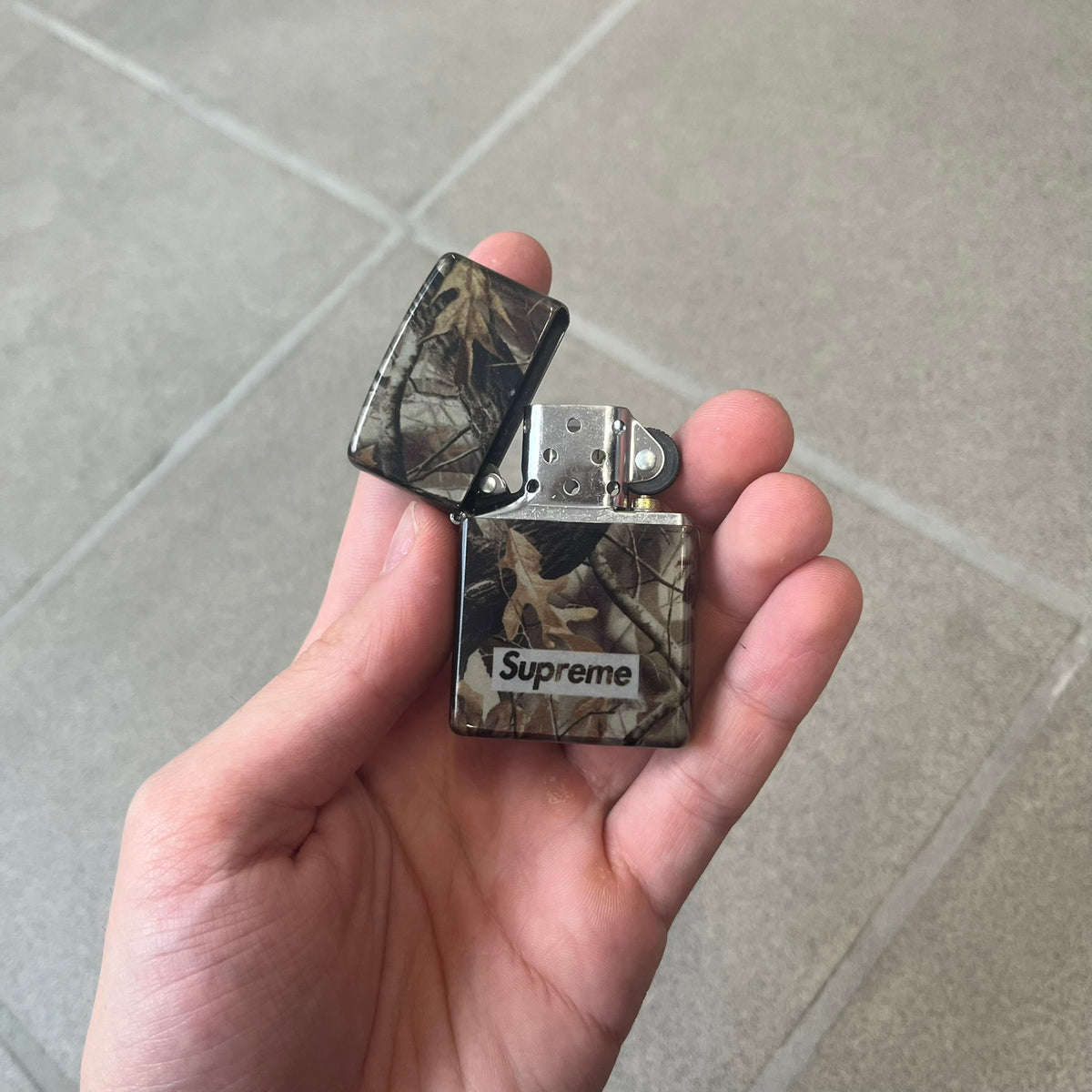 Supreme FW26 RealTree Camo Logo Zippo Lighter is an original Zippo featuring a full RealTree camo graphic with the Supreme box logo on the front. It comes with the original Zippo box and has never been filled.