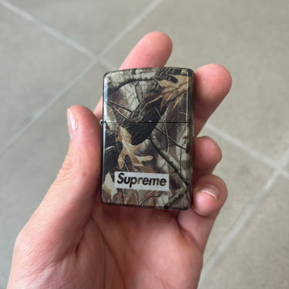 Supreme FW26 RealTree Camo Logo Zippo Lighter is an original Zippo featuring a full RealTree camo graphic with the Supreme box logo on the front. It comes with the original Zippo box and has never been filled.