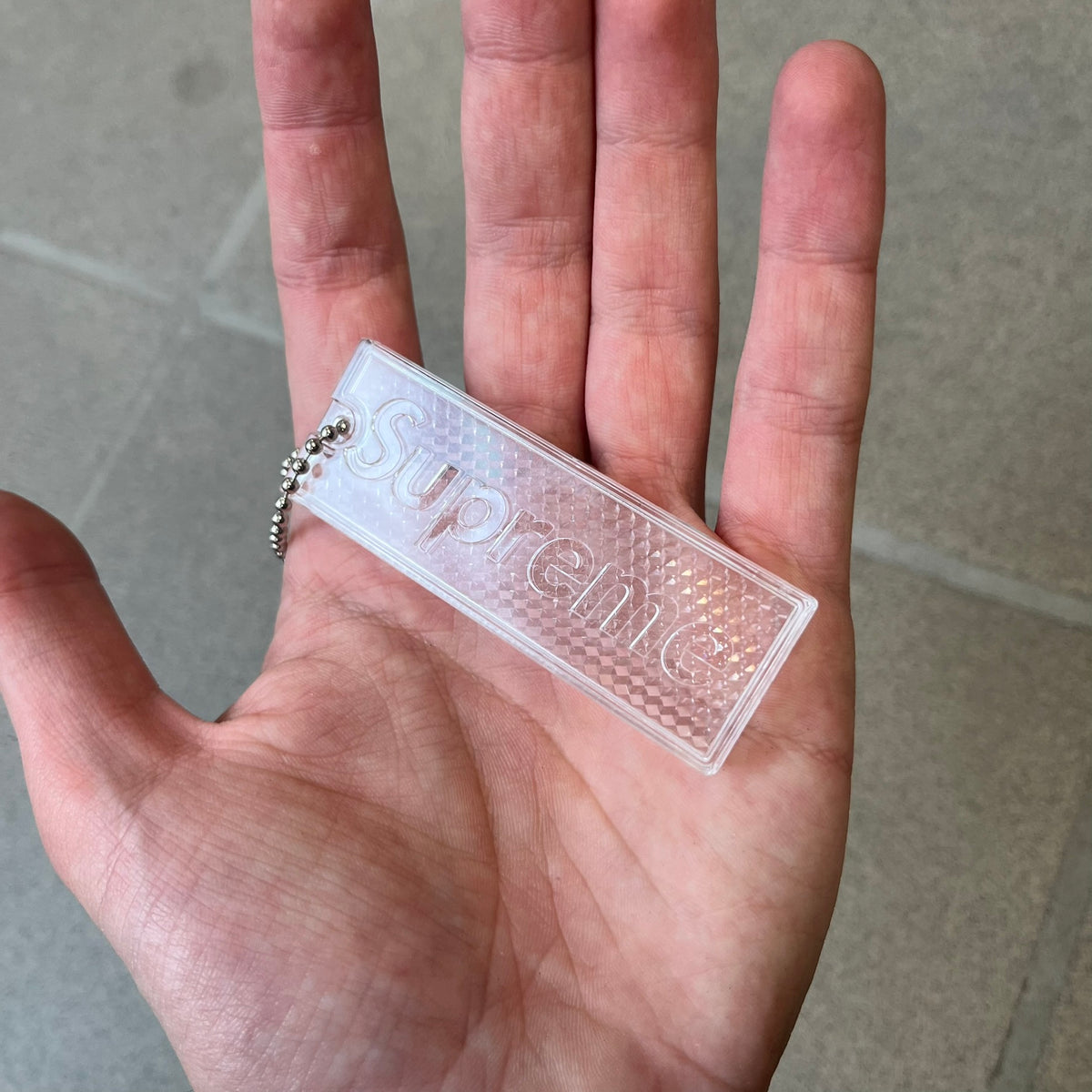 Supreme FW26 Reflective Clear Keychain is a clear plastic keychain on a steel ball chain, with a Supreme logo on the front. The back is made of small reflective panels.