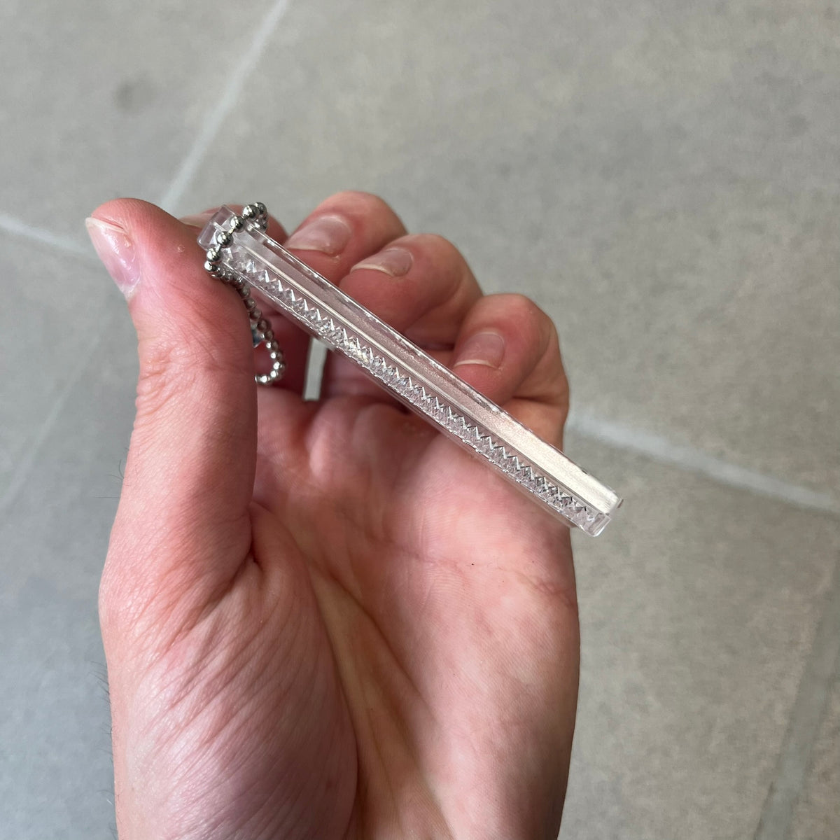 Supreme FW26 Reflective Clear Keychain is a clear plastic keychain on a steel ball chain, with a Supreme logo on the front. The back is made of small reflective panels.