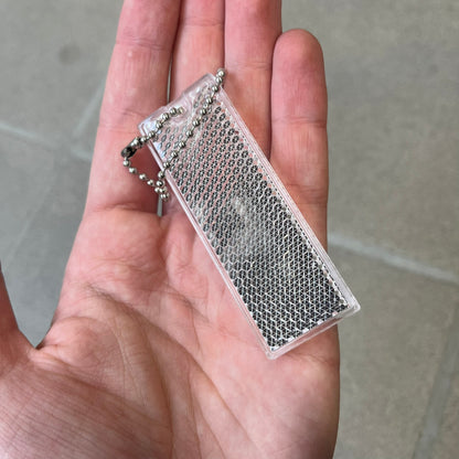 Supreme FW26 Reflective Clear Keychain is a clear plastic keychain on a steel ball chain, with a Supreme logo on the front. The back is made of small reflective panels.