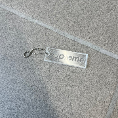 Supreme FW26 Reflective Clear Keychain is a clear plastic keychain on a steel ball chain, with a Supreme logo on the front. The back is made of small reflective panels.