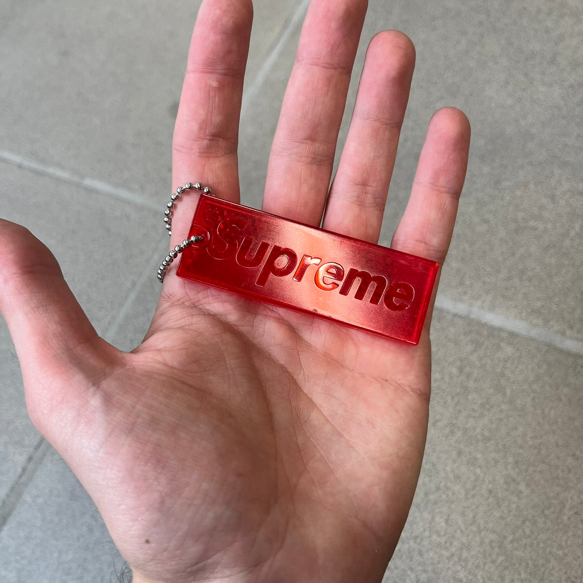 Supreme FW26 Reflective Red Keychain is a red plastic keychain on a steel ball chain, with a Supreme logo on the front. The back is made of small reflective panels.