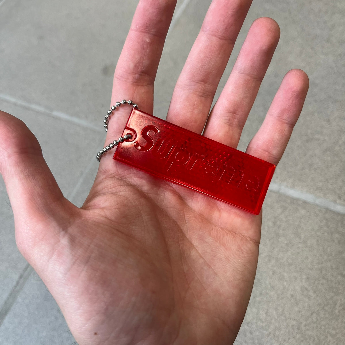 Supreme FW26 Reflective Red Keychain is a red plastic keychain on a steel ball chain, with a Supreme logo on the front. The back is made of small reflective panels.