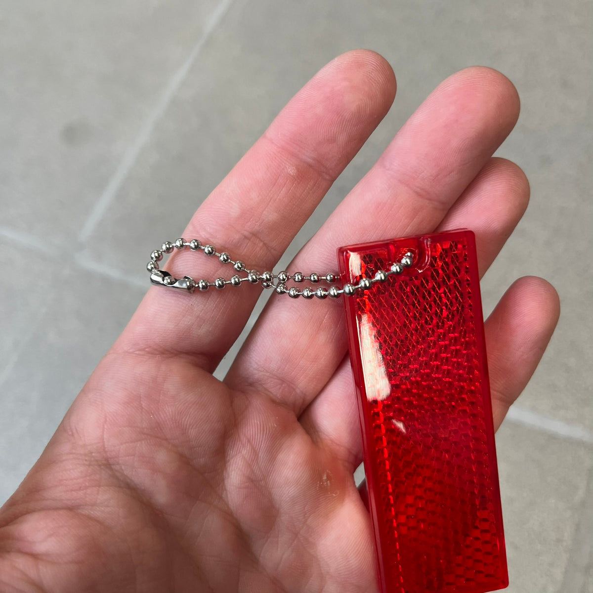 Supreme FW26 Reflective Red Keychain is a red plastic keychain on a steel ball chain, with a Supreme logo on the front. The back is made of small reflective panels.