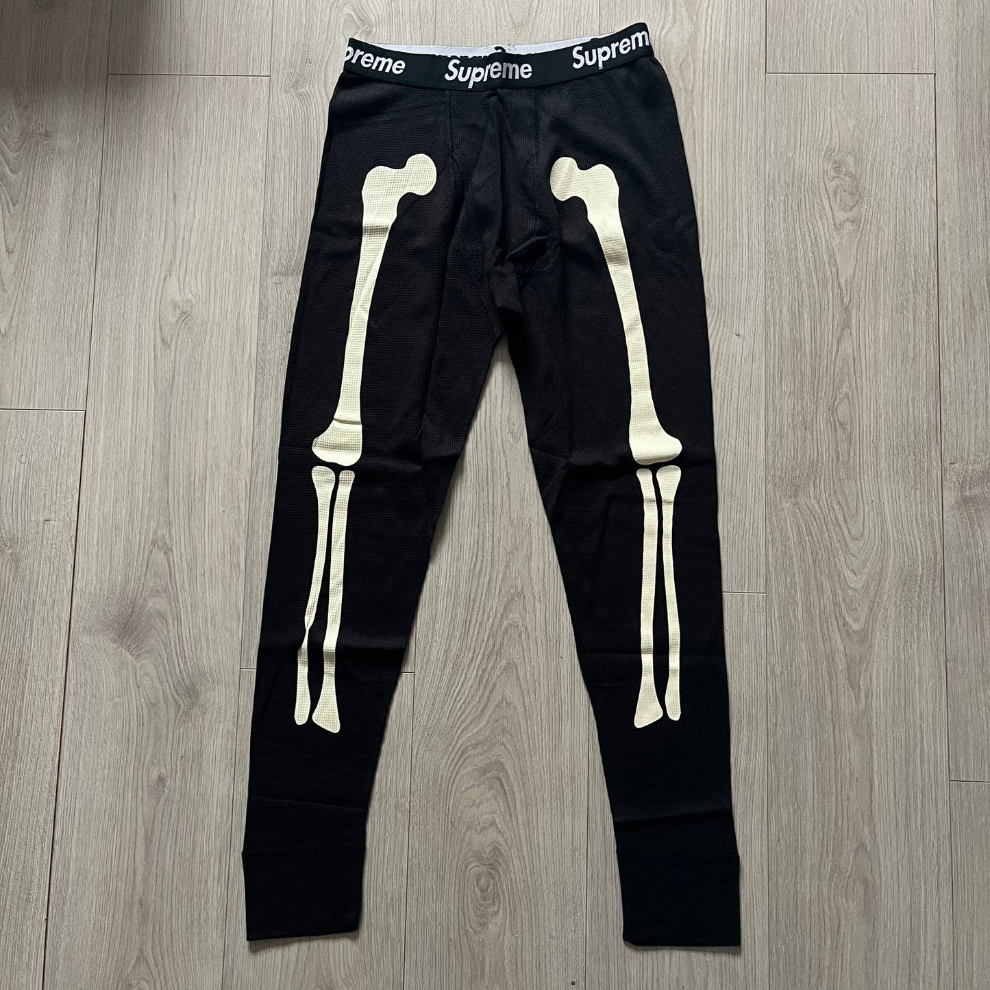  Supreme Hanes Bones Thermal Pant FW25 in black, crafted from 100% cotton. Features white skeletal bone prints running down both legs and a black elastic waistband with white Supreme branding throughout. Released October 23, 2025.