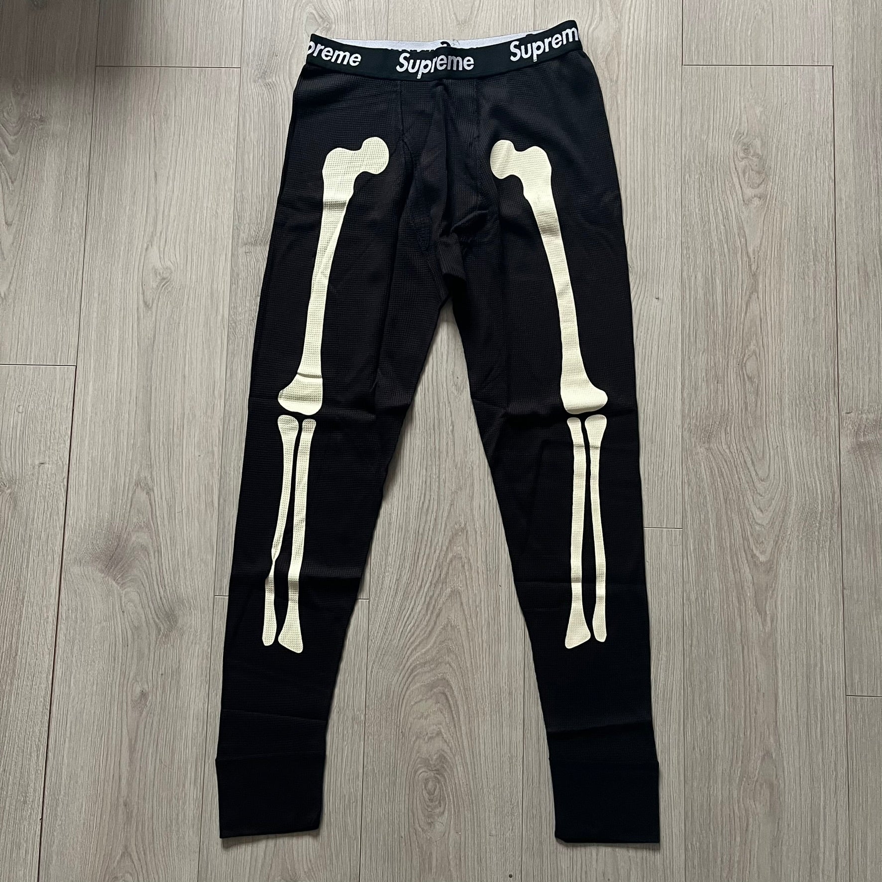  Supreme Hanes Bones Thermal Pant FW25 in black, crafted from 100% cotton. Features white skeletal bone prints running down both legs and a black elastic waistband with white Supreme branding throughout. Released October 23, 2025.