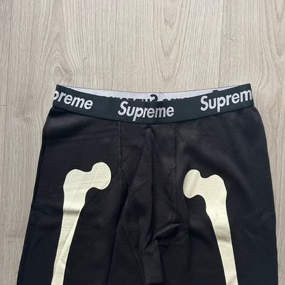  Supreme Hanes Bones Thermal Pant FW25 in black, crafted from 100% cotton. Features white skeletal bone prints running down both legs and a black elastic waistband with white Supreme branding throughout. Released October 23, 2025.